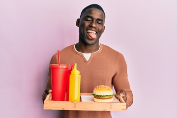 Young african american man eating a tasty classic burger and soda winking looking at the camera...