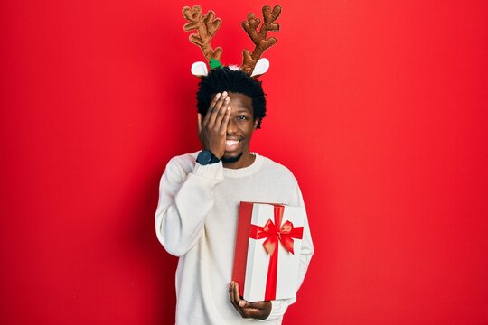 Young african american man wearing deer christmas hat holding gift covering one eye with hand, confident smile on face and surprise emotion.