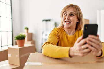 Middle age blonde woman smiling happy using smartphone at new home.