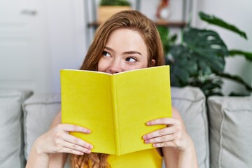 Young redhead girl smiling happy covering face with book at home