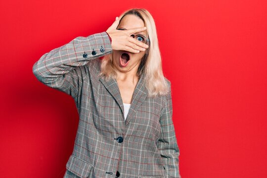 Beautiful caucasian blonde woman wearing business jacket and glasses peeking in shock covering face and eyes with hand, looking through fingers afraid