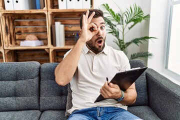 Handsome hispanic man holding clipboard working at psychology clinic doing ok gesture shocked with surprised face, eye looking through fingers. unbelieving expression.