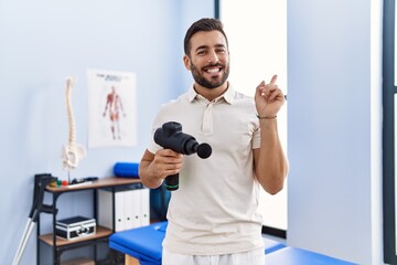 Naklejka na ściany i meble Handsome hispanic man holding therapy massage gun at physiotherapy center smiling happy pointing with hand and finger to the side