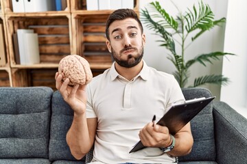 Handsome hispanic man holding brain working at psychology clinic puffing cheeks with funny face....
