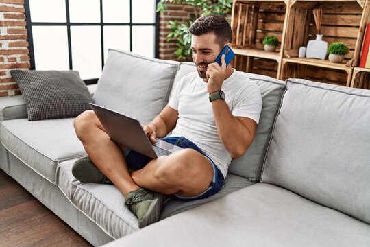 Young Hispanic Man Using Laptop And Talking On The Smartphone At Home