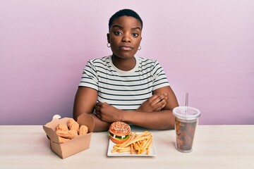 Young african american woman eating a tasty classic burger with fries and soda with serious expression on face. simple and natural looking at the camera.