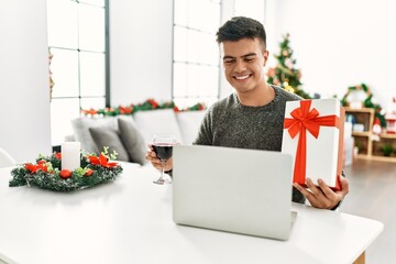Young hispanic man having video call showing gift sitting by christmas tree at home