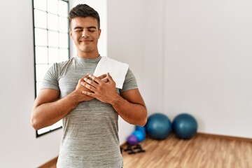 Young hispanic man wearing sportswear and towel at the gym smiling with hands on chest with closed...