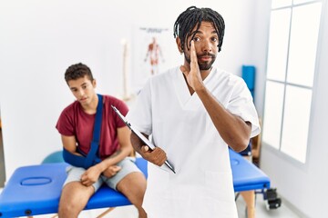 Young hispanic man working at pain recovery clinic with a man with broken arm hand on mouth telling secret rumor, whispering malicious talk conversation