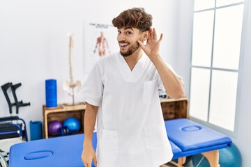 Young arab man working at pain recovery clinic smiling with hand over ear listening an hearing to rumor or gossip. deafness concept.