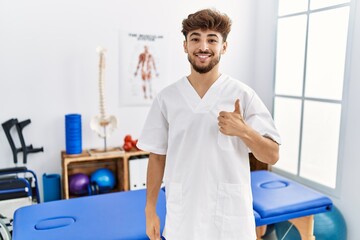 Young arab man working at pain recovery clinic doing happy thumbs up gesture with hand. approving expression looking at the camera showing success.