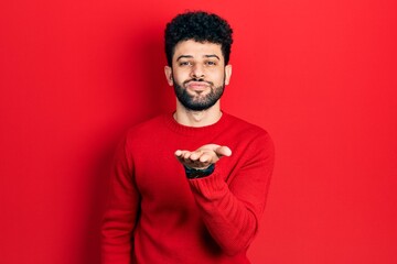 Young arab man with beard wearing casual red sweater looking at the camera blowing a kiss with hand on air being lovely and sexy. love expression.