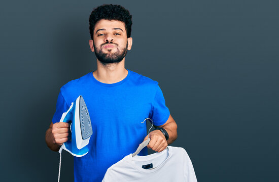 Young arab man with beard holding electric steam iron and white t shirt puffing cheeks with funny face. mouth inflated with air, catching air.