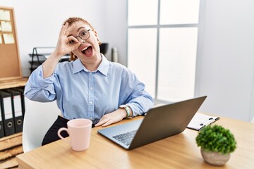 Young redhead woman working at the office using computer laptop doing ok gesture with hand smiling, eye looking through fingers with happy face.