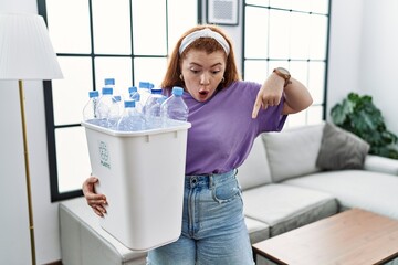 Young redhead woman holding recycling wastebasket with plastic bottles pointing down with fingers showing advertisement, surprised face and open mouth