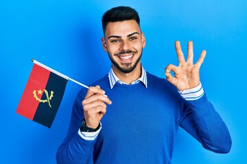 Young hispanic man with beard holding angola flag doing ok sign with fingers, smiling friendly...