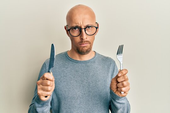 Bald man with beard holding fork and knife ready to eat skeptic and nervous, frowning upset because of problem. negative person.