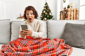 Middle age hispanic woman using smartphone sitting on the sofa by christmas tree scared and amazed with open mouth for surprise, disbelief face