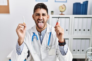 Young doctor man holding electronic cigarette at medical clinic sticking tongue out happy with funny expression.