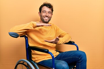 Handsome man with beard sitting on wheelchair gesturing with hands showing big and large size sign, measure symbol. smiling looking at the camera. measuring concept.