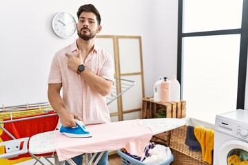 Young man with beard ironing clothes at home pointing with hand finger to the side showing advertisement, serious and calm face