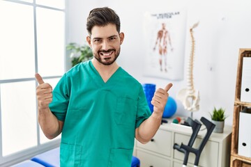 Young man with beard working at pain recovery clinic success sign doing positive gesture with hand, thumbs up smiling and happy. cheerful expression and winner gesture.