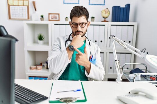 Young man with beard wearing doctor uniform and stethoscope at the clinic in hurry pointing to watch time, impatience, looking at the camera with relaxed expression