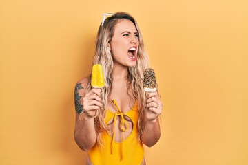 Beautiful young blonde woman wearing summer swimsuit holding ice creams angry and mad screaming frustrated and furious, shouting with anger. rage and aggressive concept.