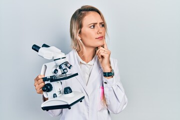 Beautiful young blonde woman holding microscope serious face thinking about question with hand on...