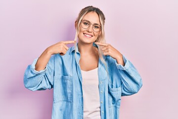 Beautiful young blonde woman wearing casual clothes and glasses smiling cheerful showing and pointing with fingers teeth and mouth. dental health concept.