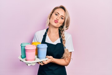 Beautiful young blonde woman wearing waitress apron holding take away cup of coffees looking at the camera blowing a kiss being lovely and sexy. love expression.