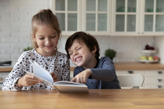 Happy little children siblings reading paper book, enjoying funny story, watching colorful pictures, enjoying spending time together, sitting at table in modern kitchen, entertaining activity concept.