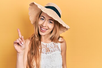 Young caucasian woman wearing summer hat showing and pointing up with finger number one while smiling confident and happy.