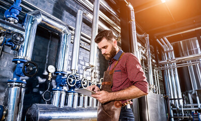Young male brewer in leather apron supervising the process of beer fermentation at modern brewery factory