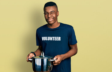 Young african american man wearing volunteer holding cooking pot winking looking at the camera with...