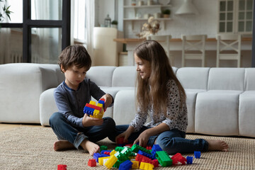 Happy adorable little brother and sister involved in creative game, playing toys sitting on floor carpet in living room. Cheerful small best friends enjoying constructing buildings with plastic cubes.