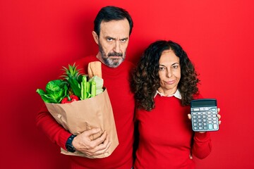 Middle age couple of hispanic woman and man holding groceries bag and calculator skeptic and...
