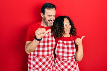 Middle age couple of hispanic woman and man wearing cook apron smiling with happy face looking and pointing to the side with thumb up.