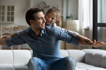 Obraz premium Joyful millennial father sitting on cozy couch, having fun playing with laughing sincere little child daughter. Happy two generations family imagining travelling, entertaining together in living room.