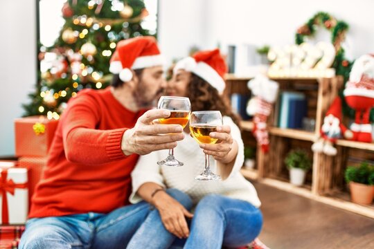 Middle age hispanic couple wearing christmas hat. Sitting on the floor toasting with champagne at home.