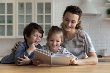 Smiling little children reading interesting story in paper book or watching pictures together with caring young mother or female babysitter, sitting at table in kitchen, hobby activity concept.