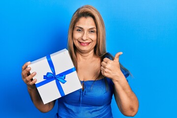 Middle age hispanic woman holding gift smiling happy and positive, thumb up doing excellent and approval sign