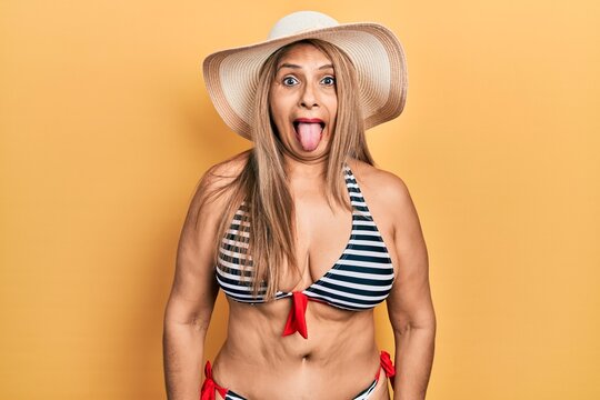 Middle age hispanic woman wearing bikini and summer hat sticking tongue out happy with funny expression. emotion concept.