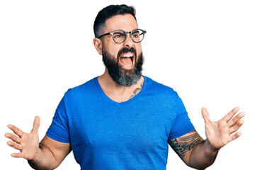 Hispanic man with beard wearing casual t shirt and glasses crazy and mad shouting and yelling with aggressive expression and arms raised. frustration concept.