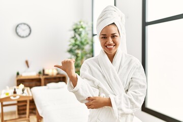 Young brunette woman wearing towel and bathrobe standing at beauty center smiling with happy face looking and pointing to the side with thumb up.