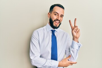 Young man with beard wearing business tie smiling with happy face winking at the camera doing victory sign. number two.