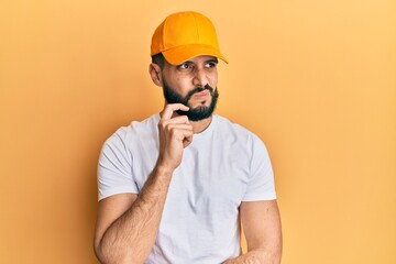Young man with beard wearing yellow cap thinking concentrated about doubt with finger on chin and looking up wondering