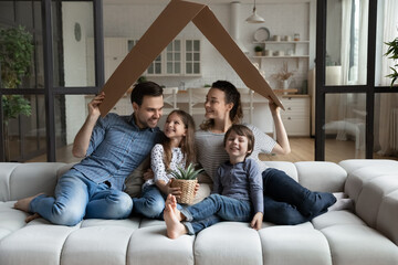 Happy bonding young couple parents and adorable little children son daughter sitting on comfortable sofa under cardboard roof, celebrating moving into new renovated apartment, real estate concept.