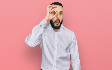 Young man with beard wearing business shirt doing ok gesture shocked with surprised face, eye looking through fingers. unbelieving expression.
