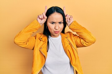 Beautiful hispanic woman with nose piercing wearing yellow leather jacket doing funny gesture with finger over head as bull horns